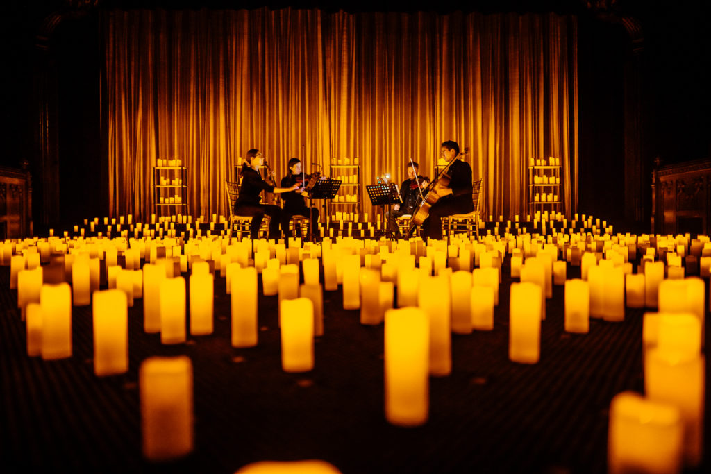 Candles cover the forefront of the image with musicians playing on a stage in the background in front of a curtain.