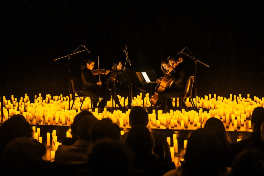 A string quartet performing on a raised stage surrounded by hundreds of candles, with a silhouette of audience members watching