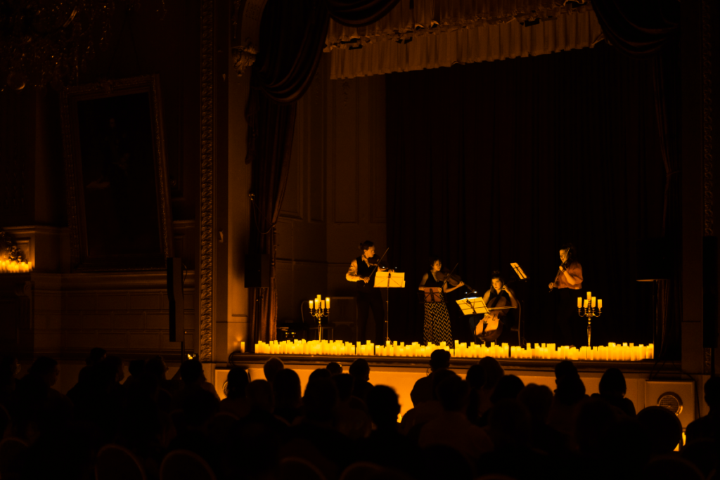A string quartet performing on stage surrounded by hundreds of candles, with the silhouette of audience members watching