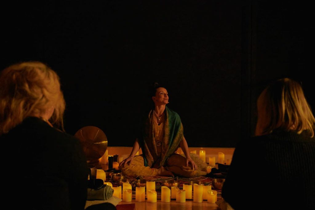 An instructor sitting in the lotus position surrounded by candles with two guests in the foreground watching her.