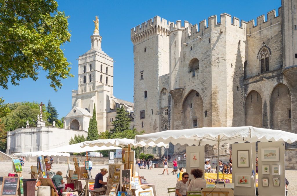 A wide shot of the Gothic Palais des Papes with a busy square full of tourists and street artists in the forefront