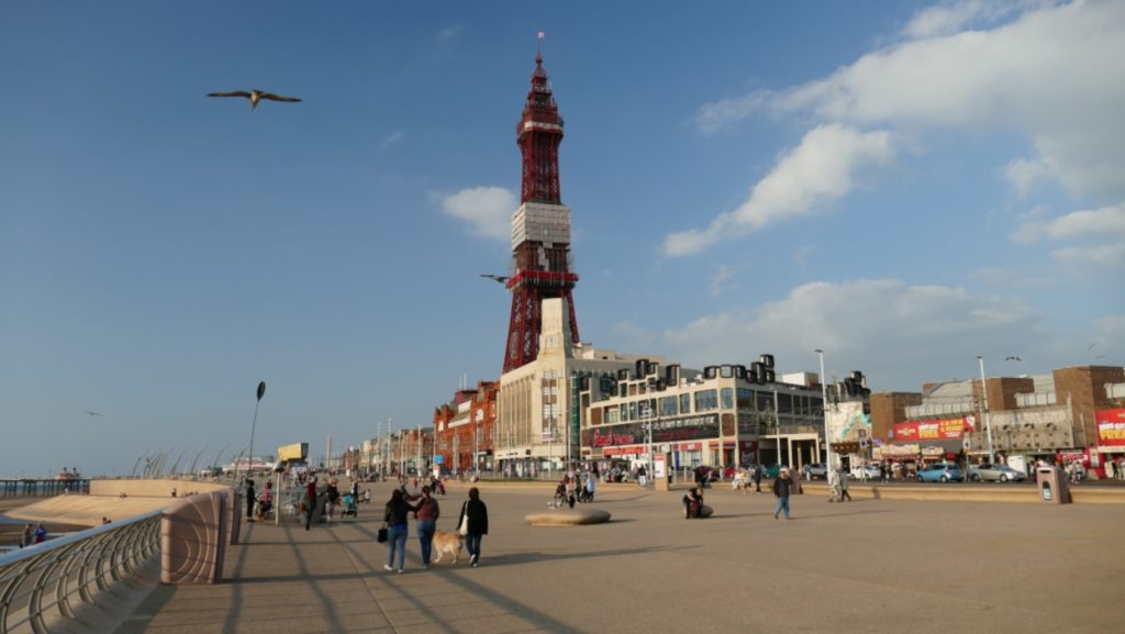 A promenade lined buy buildings in Blackpool with a blue sky and people walking along.