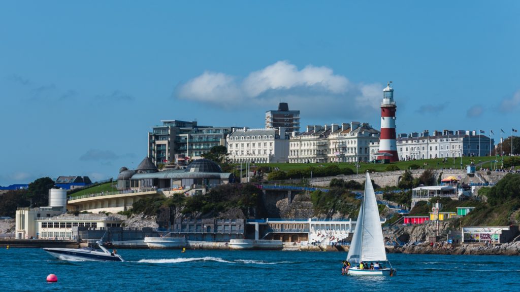 An image of Plymouth taken from the water capturing sailing boats and other boats in the channel with a lighthouse and buildings in the distance on a sunny day.