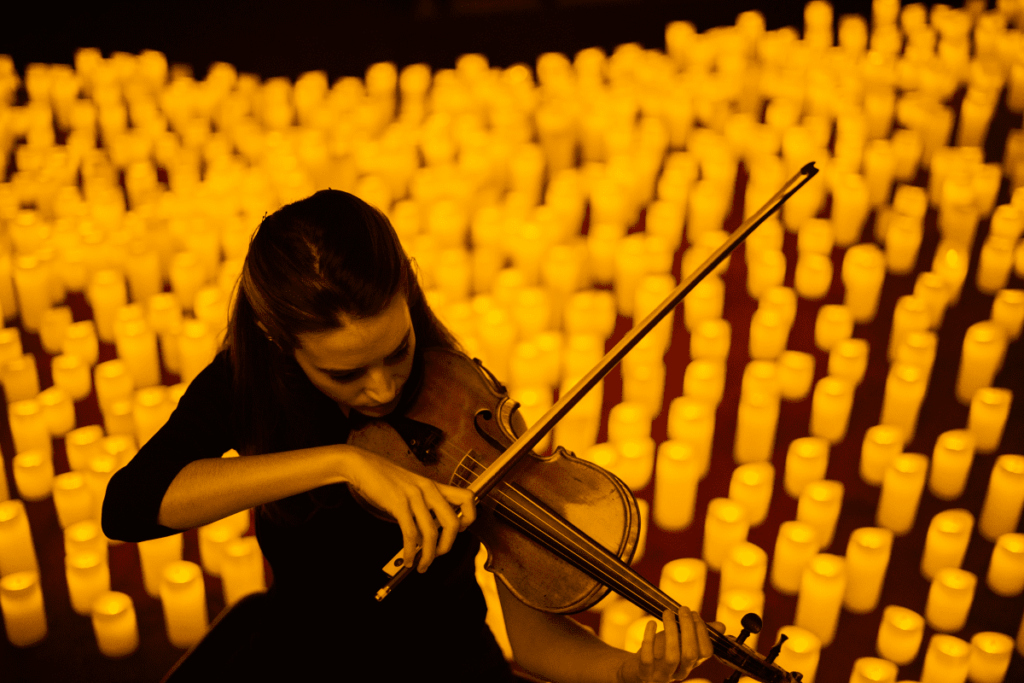 A close up of a woman playing the violin with hundreds of candles behind her.
