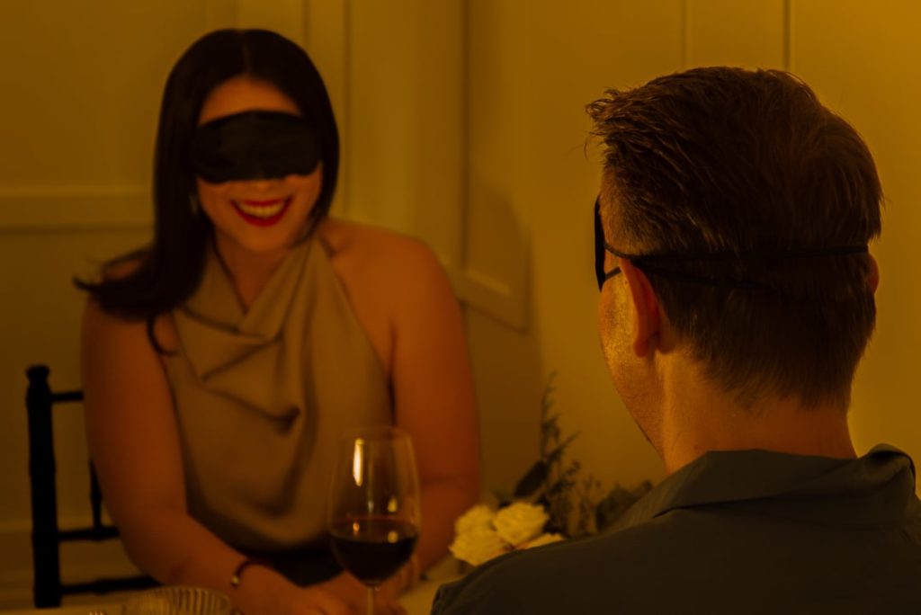A couple sit blindfolded nd smiling at Dining in the Dark.