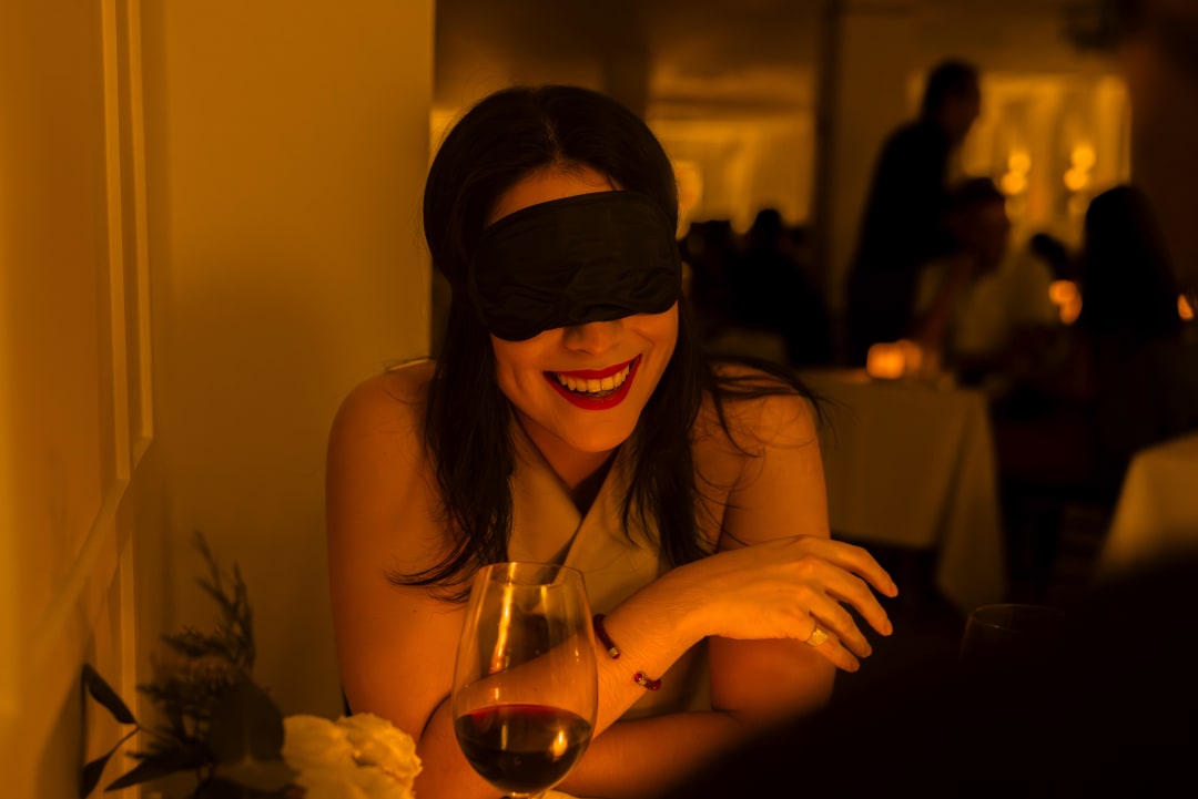 A woman laughing while blindfolded inside a restaurant with a wine glass in front of her