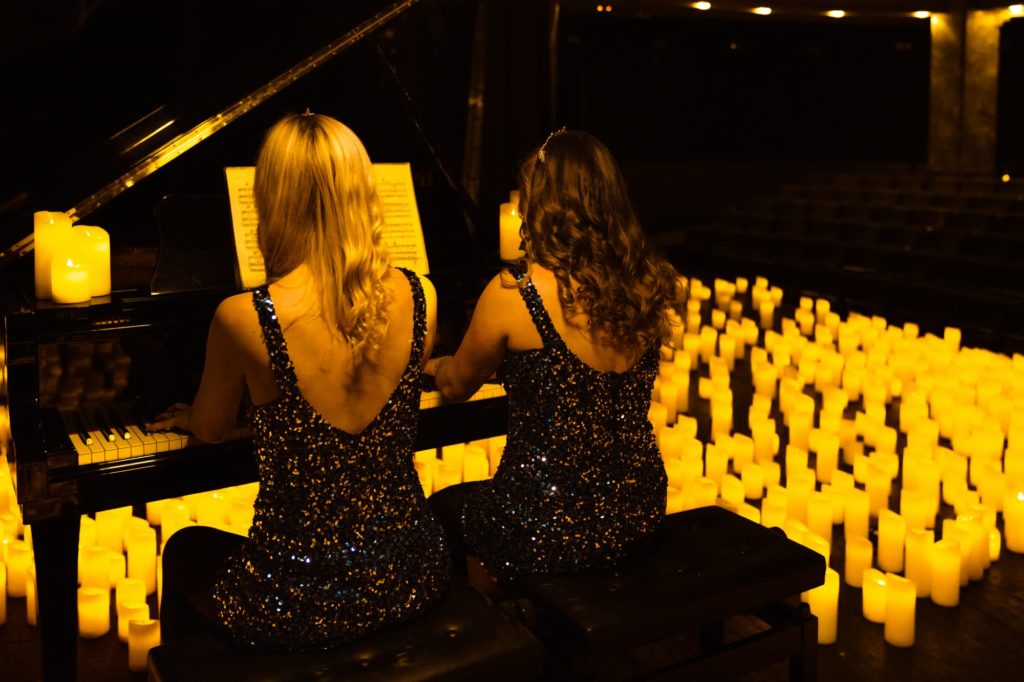 Deux femmes de dos jouent du piano à quatre mains pour un concert Candlelight entourées de bougies